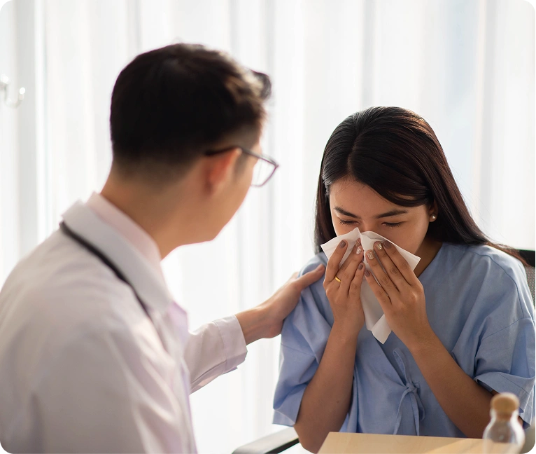 Woman sitting at a desk with a doctor standing beside her, both of them blowing their noses with a tissue