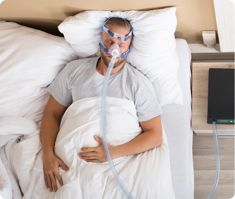 Man laying in bed with an oxygen mask on, surrounded by white pillows and a white bed sheet