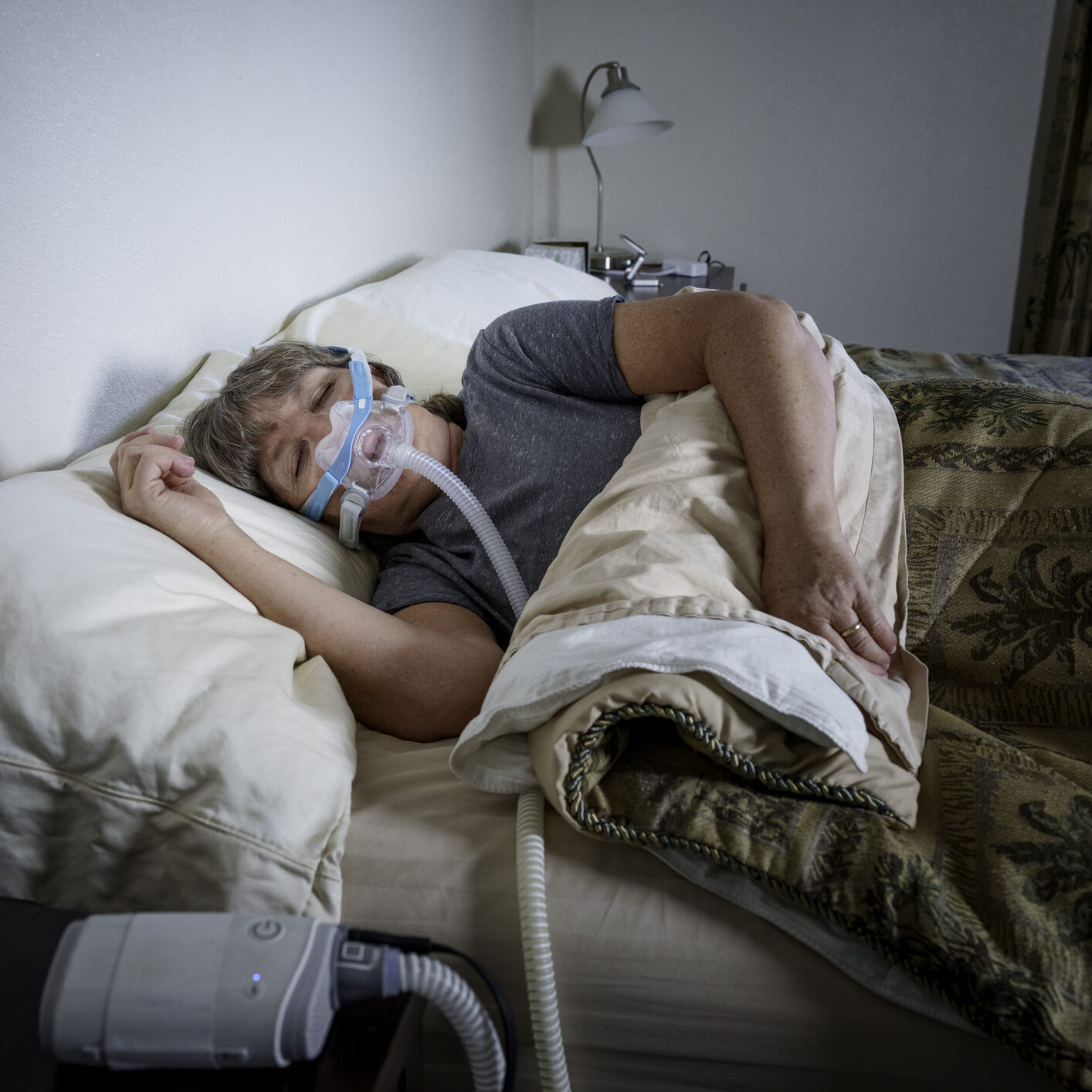 A person sleeps in bed while wearing a CPAP mask for sleep apnea treatment. The bedroom is dimly lit with a bedside lamp visible in the background. The CPAP machine sits beside the bed with tubing connected to the facial mask. The person is covered with a patterned bedspread and white pillows in a home setting. The window blinds are closed, creating a private sleeping environment.