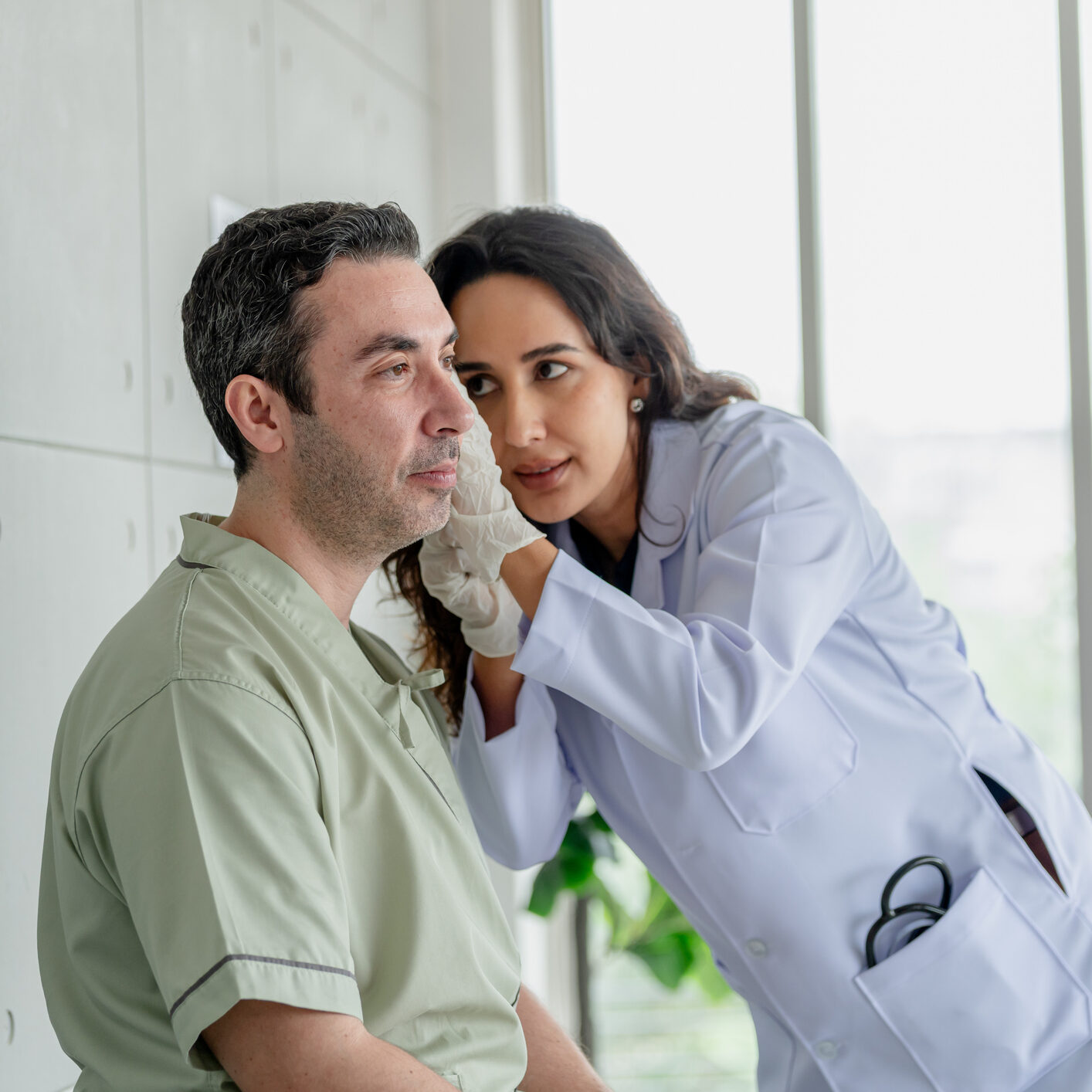 Female doctor in lab coat and surgical gloves examining ear of mature male patient in hospital. Ear examination, hearing check and medical diagnosis concept.