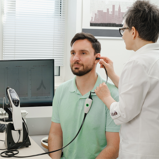 Man sitting in a chair with a stethoscope in his ear, while a woman stands beside him