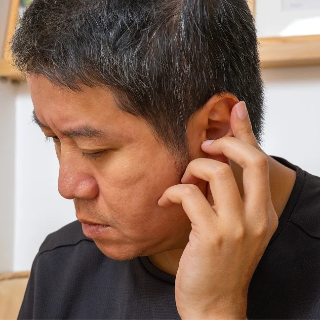 Man wearing a t-shirt, holding his ear in pain, with a frame attached to the wall in the background