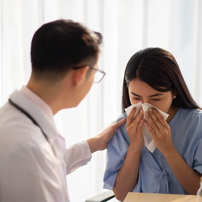Woman sitting at a desk with a doctor standing beside her, both of them blowing their noses with a tissue