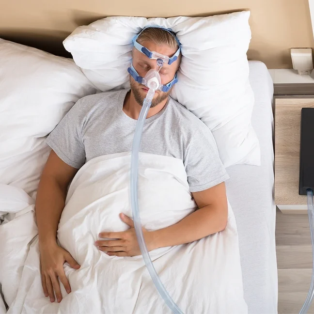 Man laying in bed with an oxygen mask on, surrounded by white pillows and a white bed sheet